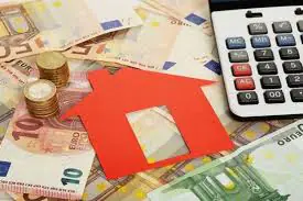 Dinero en efectivo, monedas y una calculadora junto a una figura roja de casa, simbolizando la financiación inmobiliaria o inversiones en bienes raíces.